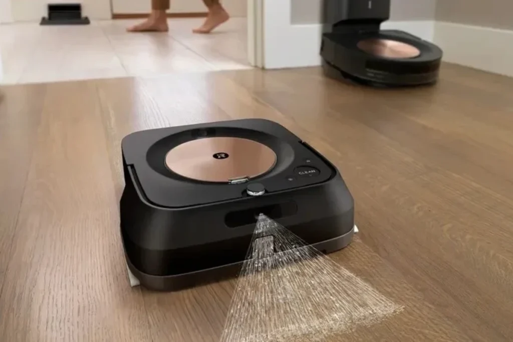 iRobot Braava M6 gulvvasker sprøjter vand på trægulv mens en person går i baggrunden og en anden robot står i døråbningen